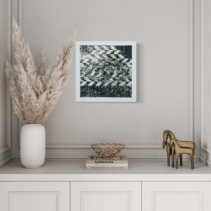 The artwork 'Waterfall' by Kiana Honarmand features an abstract geometric pattern in black and white, created from hand-cut and woven giclee prints. The 16x16 inch piece displays a cascading diagonal design reminiscent of a digital glitch or pixelated waterfall, set in a clean white frame. The composition creates a dynamic visual movement through contrasting tones and precise geometric arrangements.