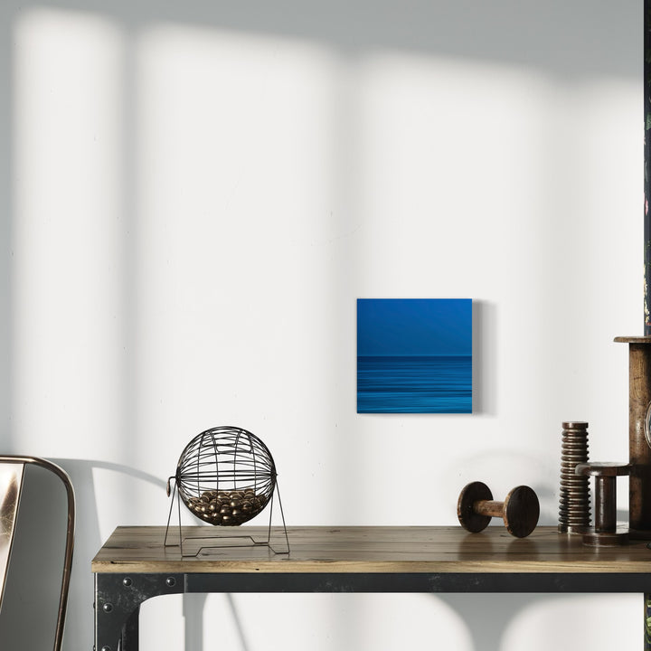 The artwork 'Blue Pacific' by Neil Shapiro features a minimalist abstract composition with horizontal bands of deep ocean blues on textured Hahnemuhle paper. The artwork creates a serene seascape impression through layered stripes, displayed against a white wall alongside industrial decor elements, capturing the essence of marine tranquility in a contemporary style.