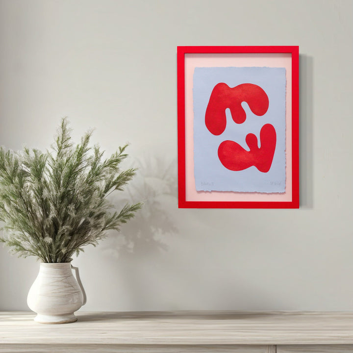 The artwork 'Balance I' by Shadia Sabagh features two abstract red organic shapes against a soft blue background, framed in vibrant red. The mirror-like forms create a dynamic composition on handmade cotton paper, painted in acrylic with a minimalist style, showcasing balance and symmetry in contemporary abstract art.