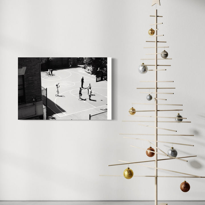 The artwork 'Ball is Life' by Cavier Coleman showcases a black and white aerial photograph of people on a basketball court, displayed on a white wall beside a minimalist wooden Christmas tree decorated with metallic ornaments. The composition captures the raw energy of street basketball in high contrast monochromatic tones, printed in 24x36 inch format.