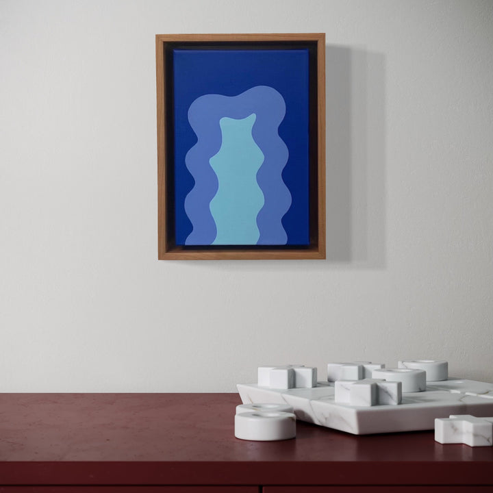 The artwork 'Blue Mini' by Shadia Sabagh features abstract wavy forms in varying shades of blue, with a lighter turquoise central element creating a fluid, vertical silhouette against a deep blue background, framed in natural wood, painted in acrylic on canvas, measuring 8x6 inches.