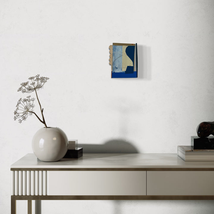 Box Top no. 64 by Janice La Motta: A minimalist collage artwork featuring abstract geometric shapes in deep blue and pale blue tones, mounted on a small 5.5 x 4.5 inch frame. The piece is displayed on a white wall above a modern console table with a spherical vase containing dried botanicals.