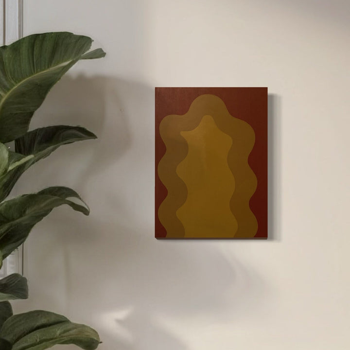 The artwork 'Brown' by Shadia Sabagh features an abstract wavy form in rich ochre and deep brown tones, creating an organic flowing pattern against a reddish-brown canvas background. Acrylic painting showcases smooth gradients and undulating lines, displayed alongside lush green foliage. Wall art measures 24x18 inches. Energy of Color Series by Shadia Sabagh.