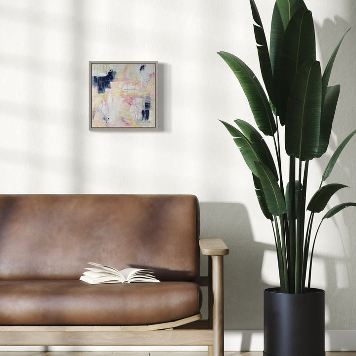 The artwork 'Buzzing Around' by Giliane Bader features abstract expressionist strokes in soft pastel pink and navy blue tones, displayed in a modern silver frame. The mixed media on birch panel with cold wax finish creates subtle textural elements, spanning 12x12 inches. Shown in a contemporary living space beside a leather sofa and tropical plant. By Giliane Bader.