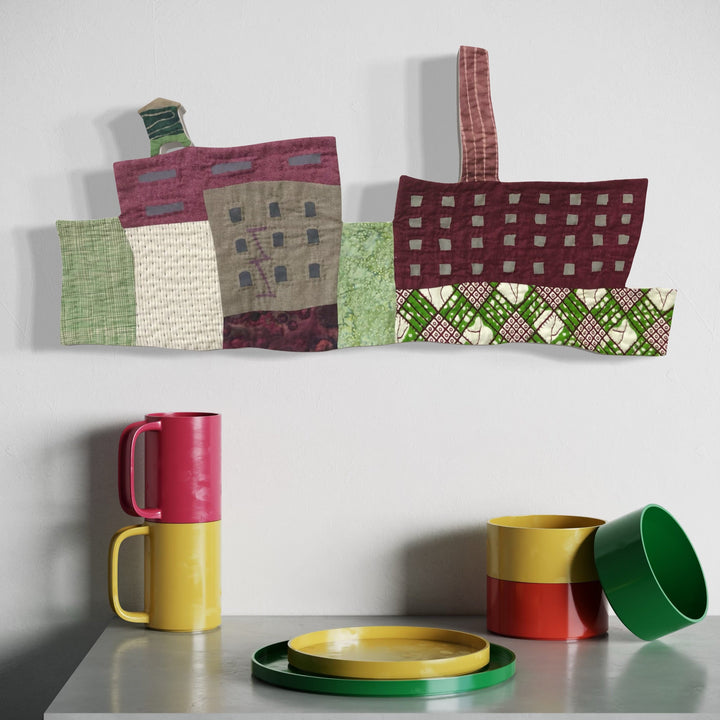 The artwork 'Cityscape (Shades of Green and Plum)' features an abstract urban landscape rendered in fabric quilting, combining geometric shapes in deep plum and varied green tones. Hand-quilted textile buildings with textured patterns create a modernist architectural composition against a white background, displaying expert craftsmanship by Ann Cofta.