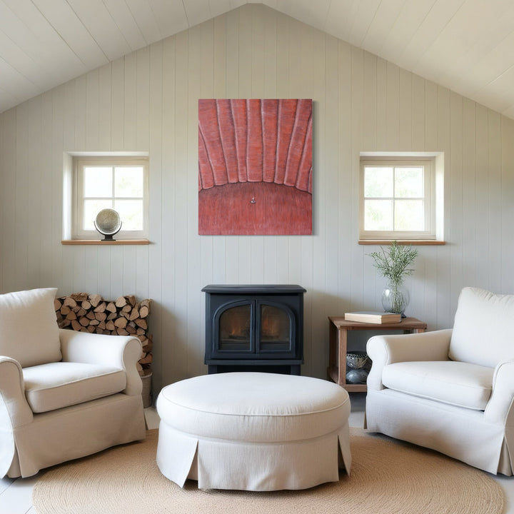 The artwork 'Comfort Zone' by Anastasia Korsakova features a minimalist living space with a striking coral-red textured canvas centered above a black wood stove. Two white slipcovered armchairs and a round ottoman create a symmetrical seating arrangement, flanked by twin windows. Natural elements include stacked firewood and a delicate plant in a glass vase.