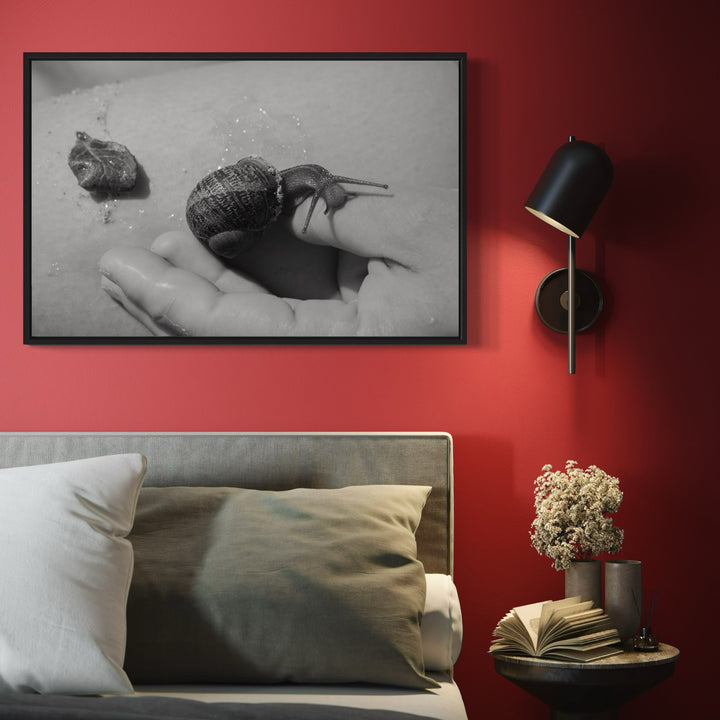 The artwork 'Communion IV' by Eunice Pais shows a striking black and white photograph of a snail crawling across an outstretched palm, creating an intimate dialogue between human and nature. The monochromatic composition emphasizes texture and form, with the snail's spiral shell casting subtle shadows. The minimalist framed photograph hangs on a bold red wall. Photo print by Eunice Pais.