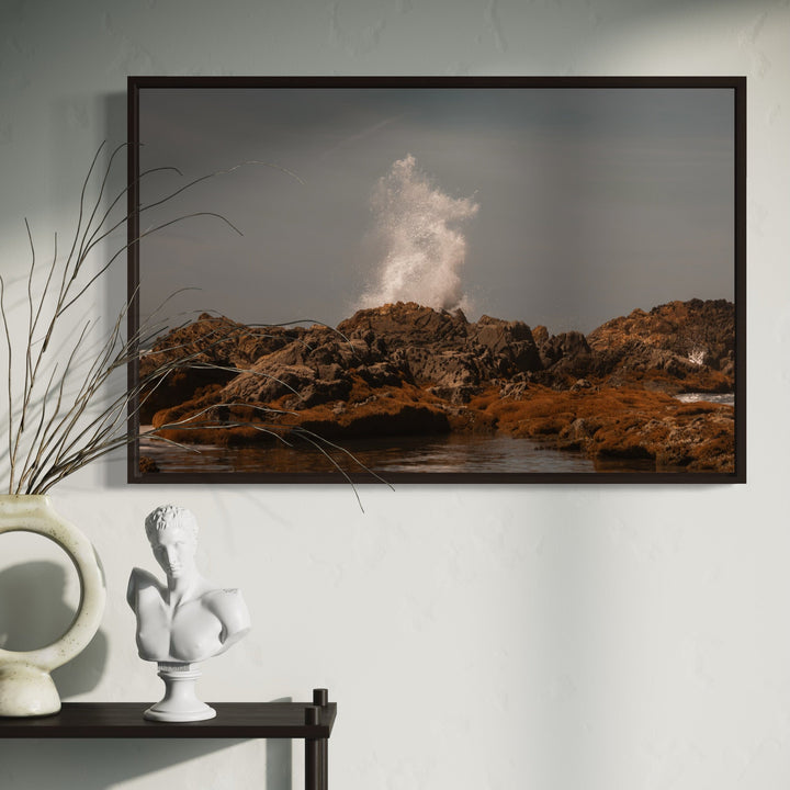 The artwork 'Communion V' by Eunice Pais captures a dramatic ocean wave crashing against rugged brown coastal rocks, creating a misty white spray against a moody gray sky. The photographic composition showcases the raw power of nature, with textured rocky formations in warm earth tones contrasting against the dynamic water movement, framed in a contemporary black border.