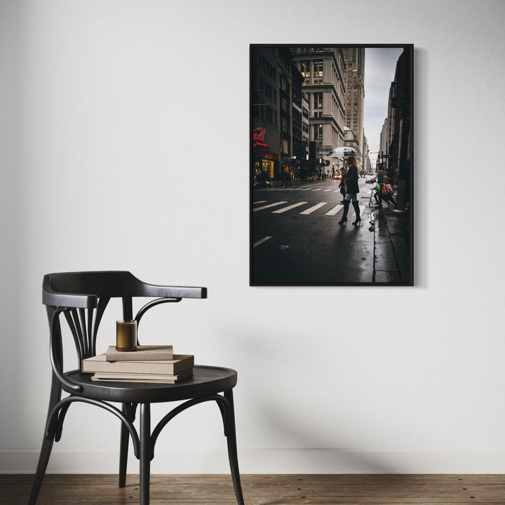 The artwork 'Daily Commute' by Dennis Maida captures a moody urban street scene in black and white photography. A silhouetted figure crosses a rain-slicked crosswalk against historic architecture, ornate building facades, and distant city structures. The dramatic contrast between light and shadow creates a cinematic atmosphere in this 36x24 inch photograph.