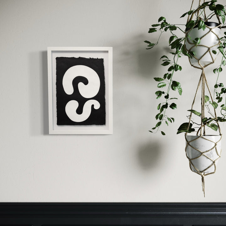 The artwork 'Duality III' by Shadia Sabagh features bold black and white abstract swirls on handmade cotton paper, displayed in a white frame against a neutral wall. The minimalist design shows curving forms creating a striking contrast, showcasing modern abstract expressionism on textured deckle-edge paper. Plant decor adds organic ambiance.