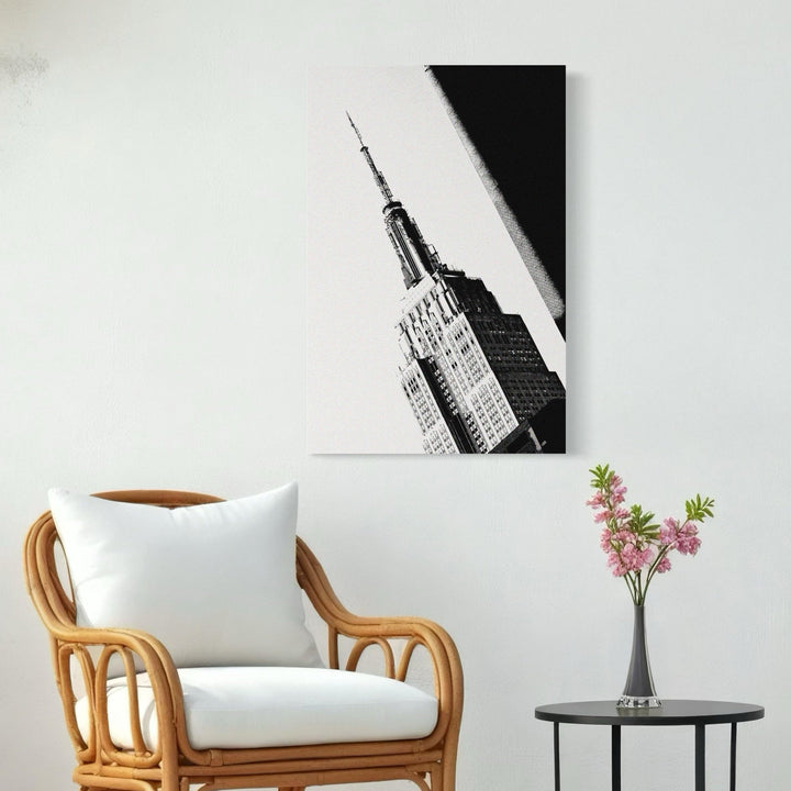 The artwork 'Empire State of Mind' captures a dramatic black and white photographic perspective of the iconic Empire State Building spire, showcasing its Art Deco architecture against a stark white background. The striking diagonal composition emphasizes the building's towering presence and geometric details. By Cavier Coleman.