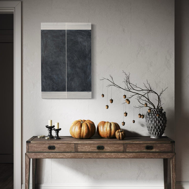 The artwork 'Erasure 34' by Erika Mahr, displayed above a rustic console table with autumn decorations, features a minimalist abstract composition in charcoal and paint on panel. The artwork showcases deep charcoal tones divided by a subtle vertical white line, creating a stark contrast against light borders, exemplifying contemporary geometric abstraction with textural depth and monochromatic sophistication.