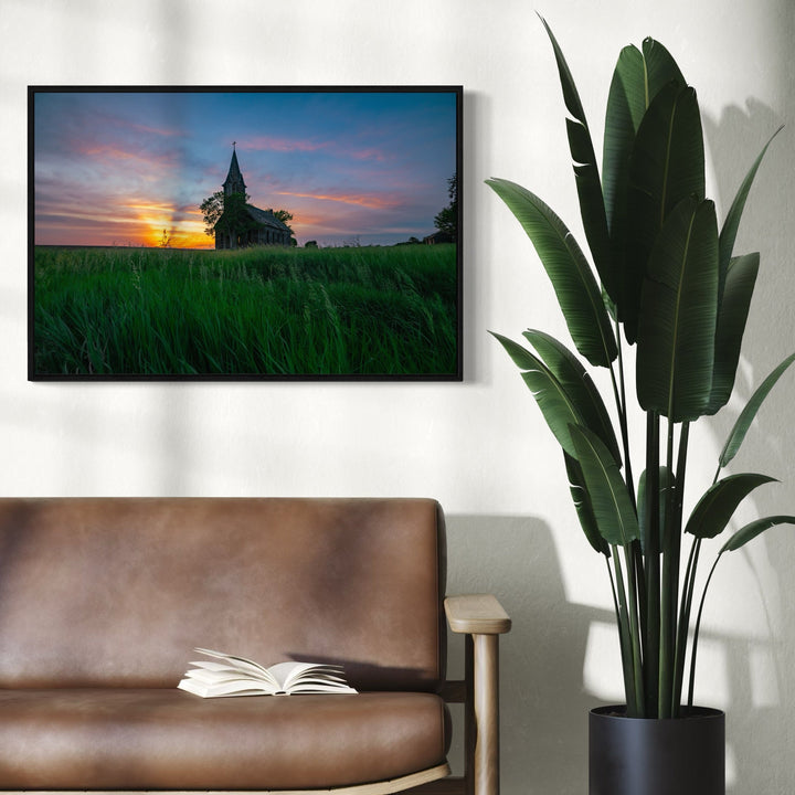 The artwork 'Evening Prayer' by Dennis Maida captures a serene rural church silhouetted against a dramatic sunset sky with vibrant orange and pink clouds. The foreground features lush green prairie grass swaying in the evening breeze, while the church's gothic spire pierces the colorful horizon, creating a peaceful and contemplative scene. Photographed on a 24x36 inch print.