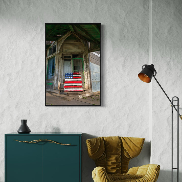 The artwork 'Faded Glory' by Dennis Maida captures a weathered wooden doorway of an abandoned building, featuring a rustic American flag painted on its entrance. The composition showcases peeling paint, distressed architecture, and muted tones of green and beige, with the vibrant red, white, and blue of the flag creating a striking focal point. The photographic art piece measures 24x36 inches.