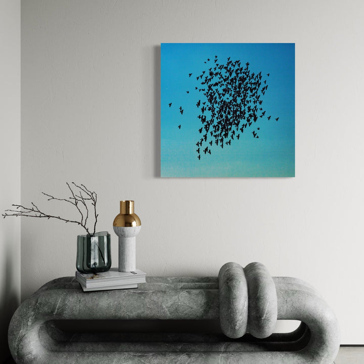 The artwork 'Flock Together' by Cavier Coleman features a striking photographic composition of silhouetted birds in flight against a vibrant turquoise sky. The birds form a dense, organic cluster floating in the upper portion of the square frame, creating a dynamic pattern of black shapes that disperses towards its edges. The minimalist composition and gradient blue background creates a serene, contemplative atmosphere. 30x30 inches photographic 