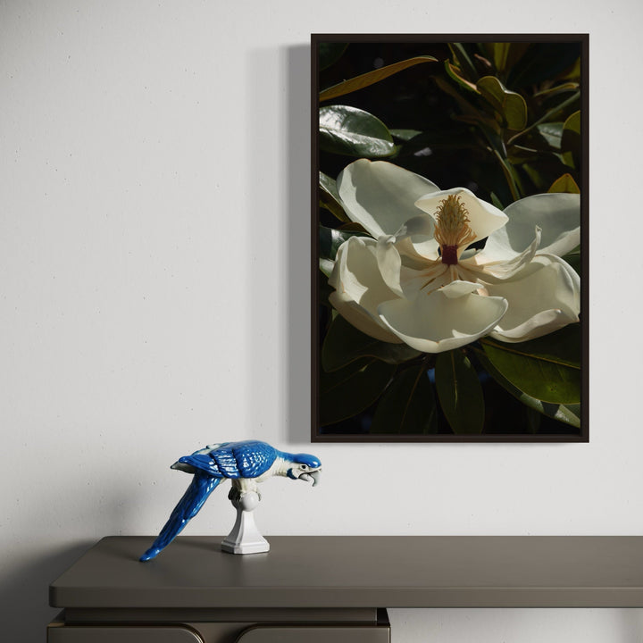 The artwork 'Flower Study III' by Eunice Pais captures a luminous white magnolia bloom in dramatic close-up, showcasing delicate petals and central pistils against dark green foliage. The photographic composition emphasizes natural light and shadow, creating depth and texture in this botanical fine art piece framed in black.