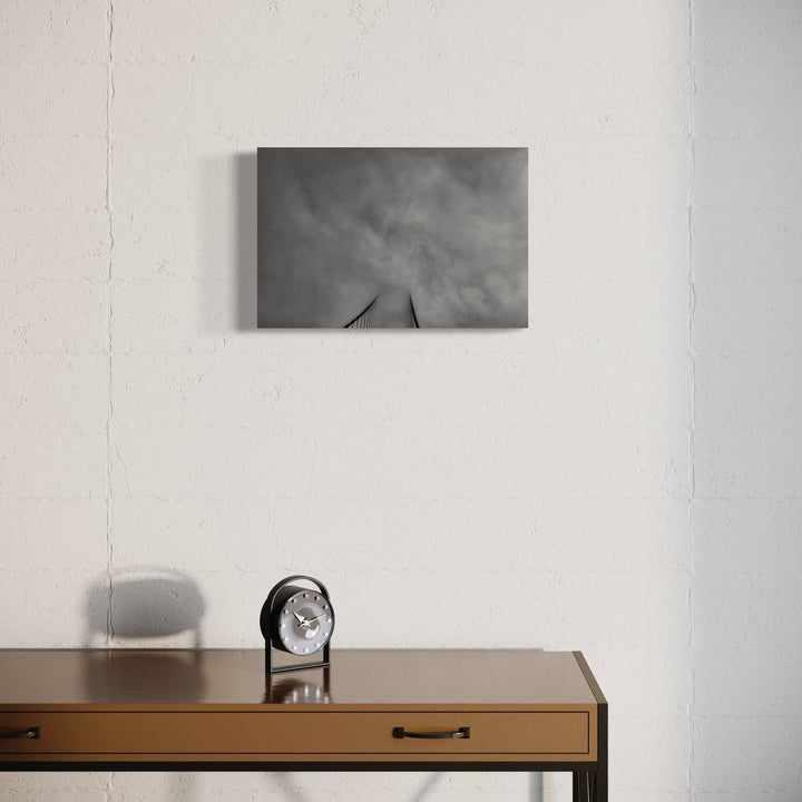 The artwork 'Golden Gate Bridge' by Neil Shapiro features a dramatic black and white photograph mounted on a white wall above a wooden desk. The minimalist composition captures the iconic bridge's towers emerging into moody, cloud-filled skies, printed on textured Hahnemühle paper for museum-quality depth and contrast.