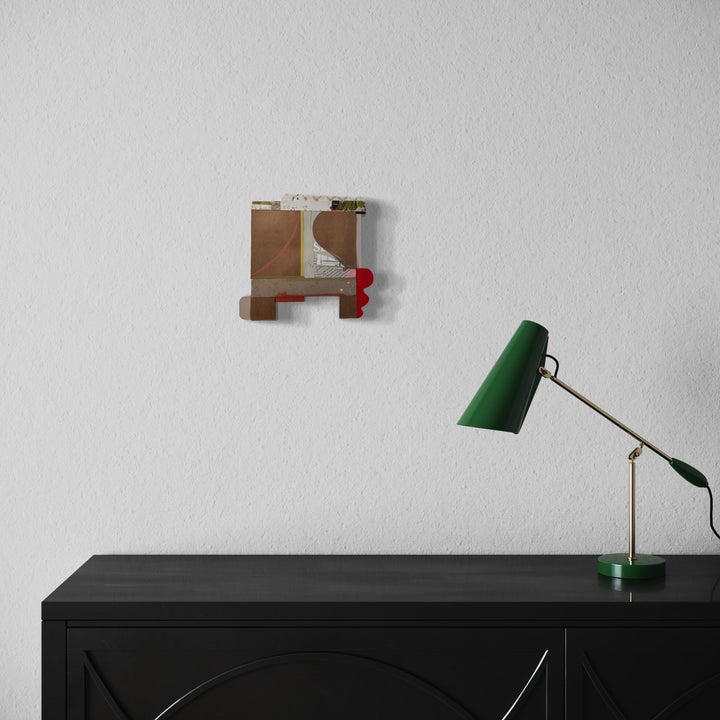 The artwork 'Good Rose' by Janice La Motta features an abstract collage composition with geometric brown paper fragments arranged in a square formation, accented by a small red detail. The minimalist piece is mounted on a white wall above a black cabinet with a vintage-style green desk lamp. 9x9.5 inches collage artwork by Janice La Motta.