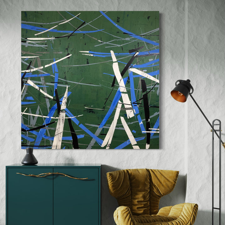 The artwork 'Green Conversation' by Shira Toren features dynamic intersecting lines in white, blue, and black against a deep forest green background. The abstract composition, created with Venetian plaster and graphite, suggests a rhythmic movement of angular shapes, resembling a modernist interpretation of branches or geometric patterns. The 50x50 inch canvas showcases textural elements and bold contrasts. By Shira Toren.