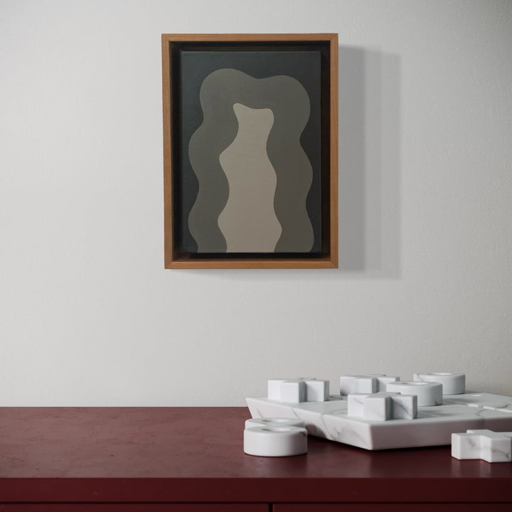 The artwork 'Grey Mini' by Shadia Sabagh features layered organic wavy shapes in varying shades of grey, creating a dimensional effect within a wooden frame. The abstract composition displays fluid curves against a dark background, showcasing depth through tonal gradation in acrylic on canvas, 8x6 inches.