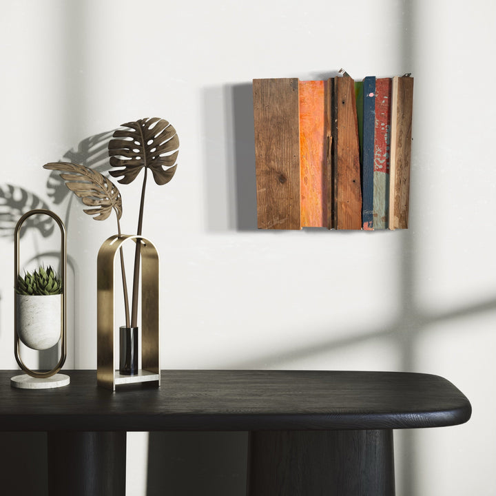 The artwork 'Griffin' by Simon Draper features weathered wooden book spines arranged horizontally on a wall, showcasing rustic textures in earth tones of brown, orange, and teal. A wall-mounted painted wood assemblage measuring 12x12 inches, capturing the nostalgic essence of vintage books through distressed materials and subtle shadows. The composition creates an illusion of actual books in a minimalist style.