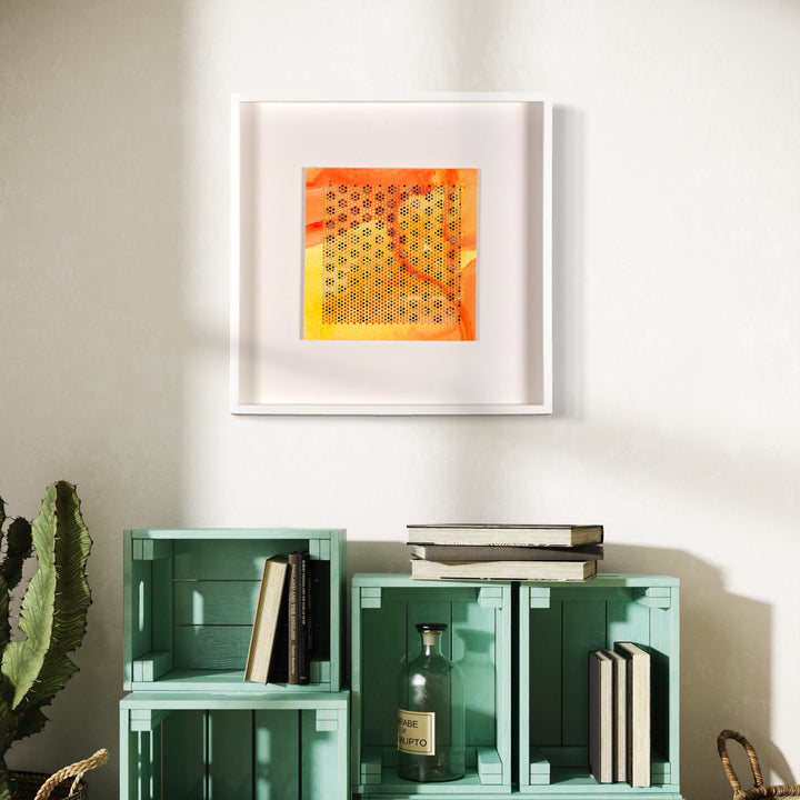 The artwork 'Hay' by Margaret Griffith features a vibrant geometric pattern of interconnected hexagonal shapes in warm orange and yellow hues, presented on cut paper within a crisp white frame. The honeycomb-like design creates a mesmerizing optical effect, showcasing precise acrylic application and meticulous paper-cutting technique. Mounted above turquoise storage crates. Original artwork by Margaret Griffith.