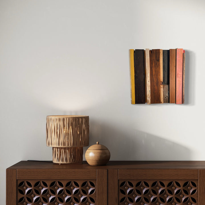 The artwork 'Jabberwock' by Simon Draper features a geometric abstract wall sculpture composed of vertical painted wood pieces in earth tones of brown, yellow, coral, and natural wood. The 12x12 inch assemblage creates a rhythmic pattern of varying heights and widths, mounted against a neutral wall above a wooden cabinet with ornate geometric cutouts.