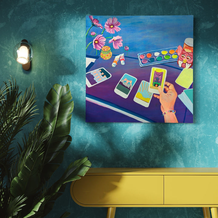 The artwork 'Justice in Her Hands' by Mary Younkin depicts a vibrant scene with tarot cards spread on a purple surface, accompanied by art supplies including a watercolor palette. Pink cosmos flowers in a yellow vase add life to the composition, set against a deep blue background. The 30x30 inch acrylic painting on wood panel features bold colors and modern storytelling elements. Art by Mary Younkin.