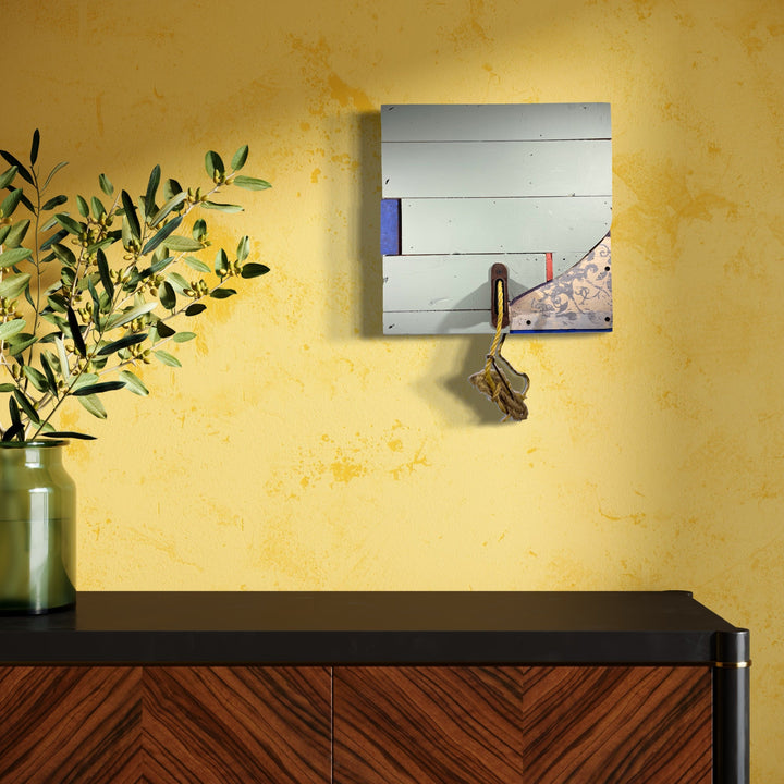 Kingston Shutter no. 30 by Simon Draper: Minimalist wall assemblage featuring horizontal white painted wood slats with subtle blue and red geometric accents, mounted on a 16x16 inch square. Small decorative hook detail against textured yellow wall, complemented by modern furniture and plant decor.