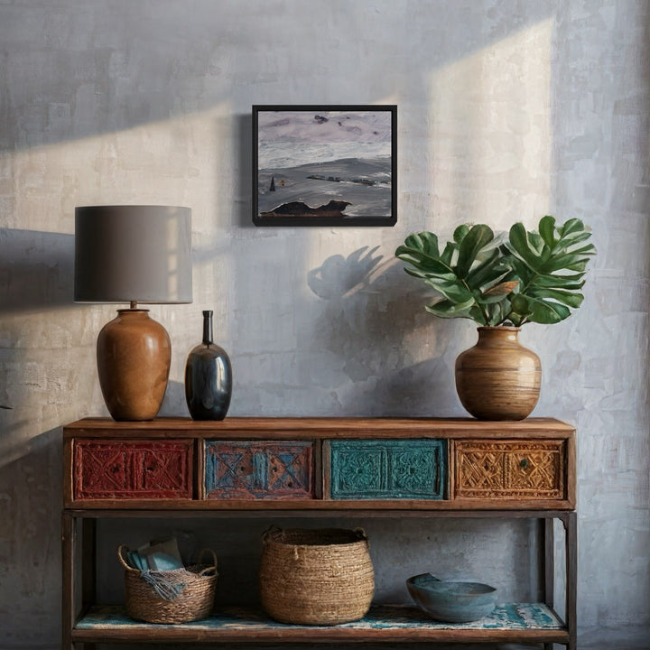 The artwork 'Landing' by Marianne Barcellona displayed above a rustic console table featuring multicolored carved drawers in red, blue, and turquoise. The mixed media piece, created with ink, acrylic, and paper collage, is shown in a black frame against a textured gray wall, accompanied by decorative elements including a terracotta lamp, monstera plant, and woven baskets.