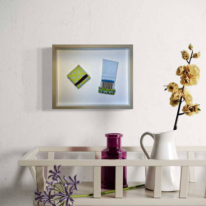 The artwork 'Matchbooks (Blue & Green)' by Ann Cofta features two floating matchbook designs on a white background in a shadow box frame. One matchbook appears in light blue with exposed matches, while the other displays a vibrant lime green pattern with yellow dots. The textile artwork combines fabric applique and paint, creating a whimsical, three-dimensional effect.