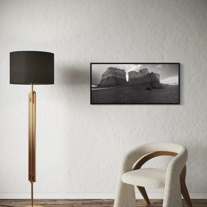 The artwork 'Monuments of Time' by Dennis Maida features a dramatic black and white panoramic photograph of towering desert rock formations. The composition captures weathered sandstone cliffs against an expansive sky, creating a stark monochromatic landscape that emphasizes the natural geological textures and erosion patterns. Framed in black, this 40x18 inch photograph showcases the timeless majesty of desert architecture.