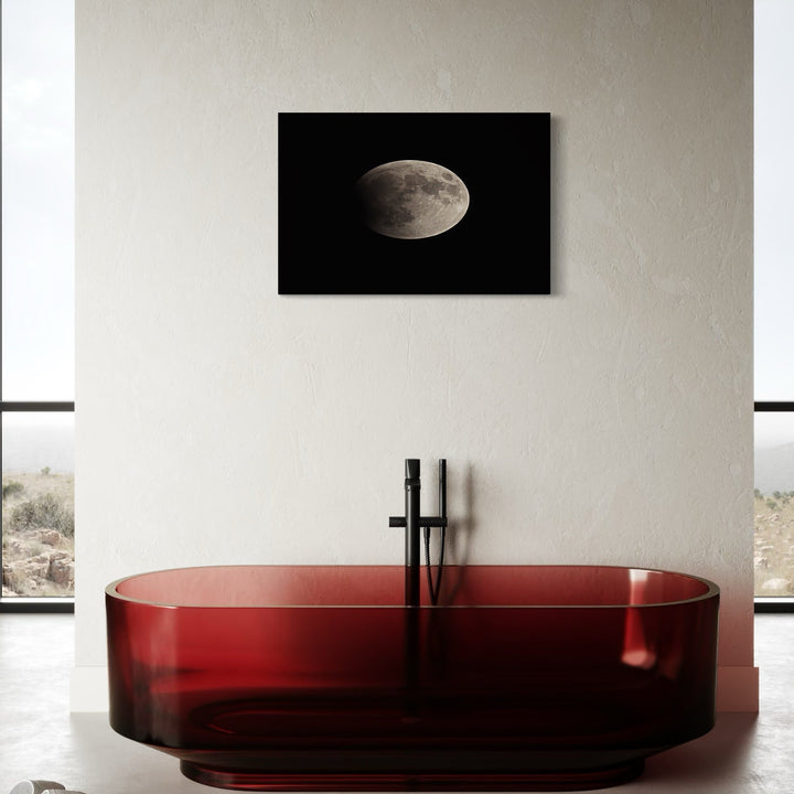 The artwork 'Moon' by Cavier Coleman features a dramatic black and white lunar photograph mounted above a modern translucent red bathtub. The moon appears in its gibbous phase against a deep black background, capturing intricate surface details. The contemporary bathroom setting showcases minimalist design with clean white walls and floor-to-ceiling windows. Size: 24x36 inches. Photography by Cavier Coleman.