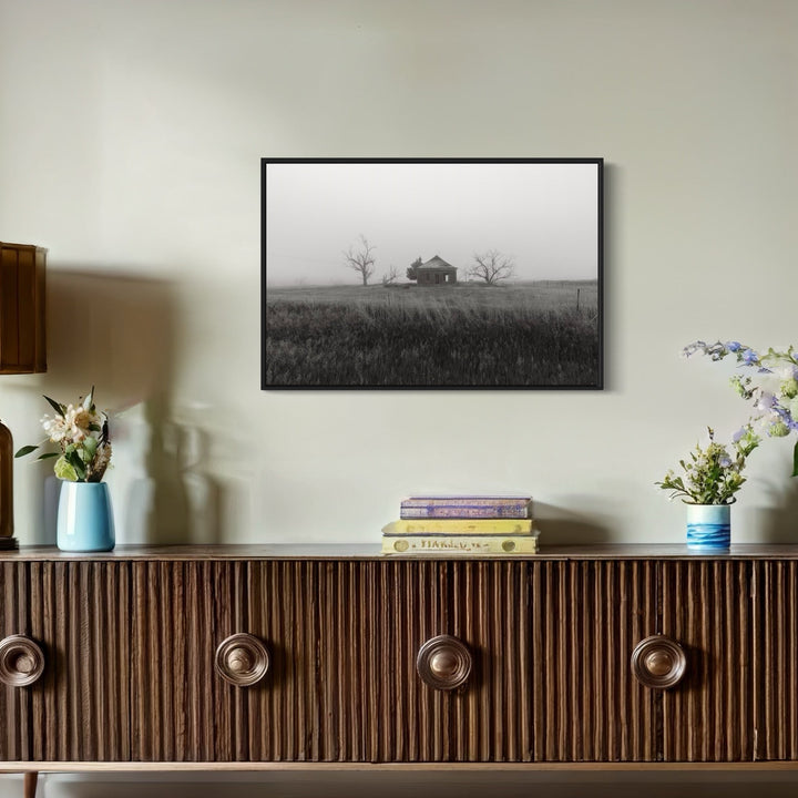 The artwork 'Nevermore' by Dennis Maida captures a haunting black and white photograph of an isolated farmhouse on a misty horizon, flanked by two bare trees. The composition features tall prairie grass in the foreground and a moody, atmospheric sky, creating a stark minimalist landscape with rich tonal contrast. Printed as a 24x36 inch photograph.