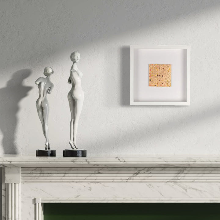 The artwork 'Ogilvy' by Margaret Griffith features a minimalist square composition in peachy-orange tones with delicate black dots scattered across the surface, displayed in a crisp white frame. The geometric abstract piece sits on a white wall above a classic marble mantelpiece, creating a striking contemporary contrast with decorative sculptural elements.