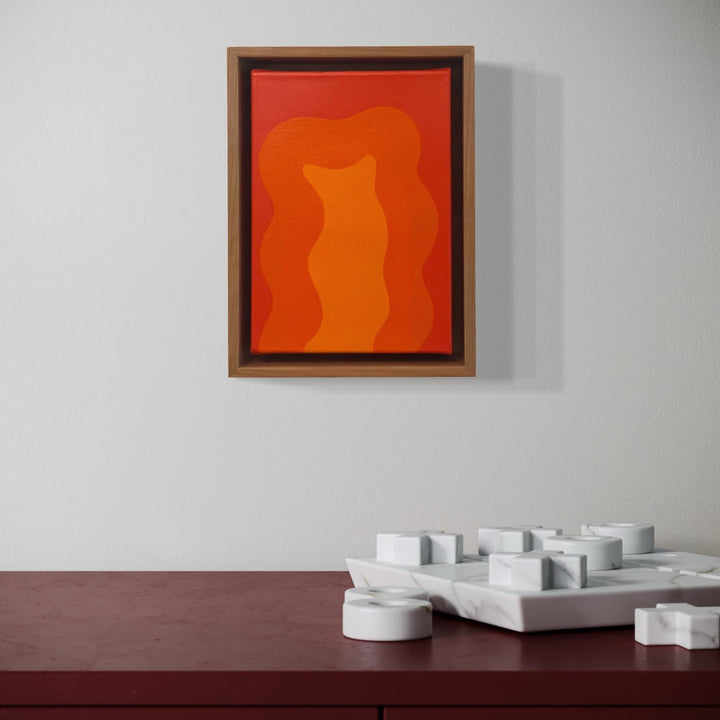 The artwork 'Orange Mini' by Shadia Sabagh features vibrant orange waves flowing organically against a darker orange background, framed in wooden brown and black borders. Small canvas acrylic painting displaying abstract minimalist style with fluid forms, mounted on white wall, measuring 8x6 inches.