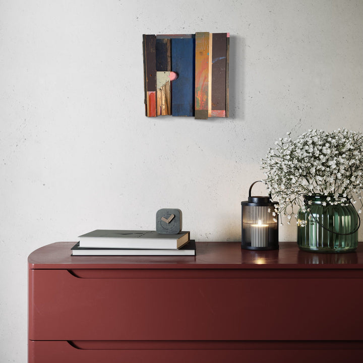 The artwork 'Prebuilt' by Simon Draper features geometric wooden assemblage with contrasting vertical panels in deep navy blue, warm brown, and coral tones. Abstract composition mounted on wall above burgundy dresser, showcasing textural painted wood elements arranged in modernist style. 12x12 inches painted wood construction with layered architectural forms.