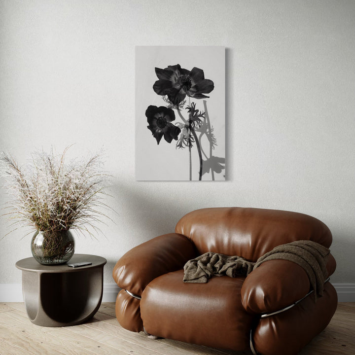 The artwork 'Reflective Beauty' by Cavier Coleman features a dramatic black and white photographic print of dark blooming flowers against a light background, mounted above a brown leather accent chair. The composition includes delicate stem details and striking floral silhouettes. A monochromatic botanical study capturing natural elegance and shadow play.