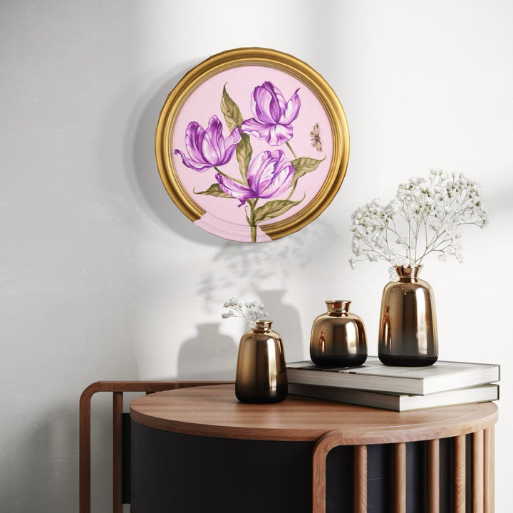 The artwork 'Rose Tulips' by Carlos Gamez de Francisco features vibrant purple tulip blooms against a soft pink background, encased in a circular gold frame. The delicate brushstrokes in acrylic capture three tulips with green stems and leaves, showcasing a contemporary botanical style on a 12x12 inch canvas.