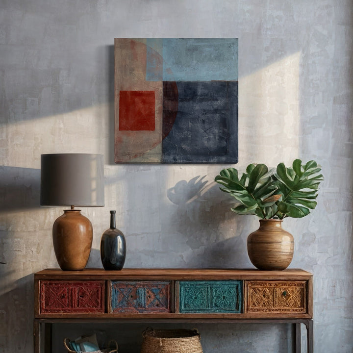 The artwork 'Rust and Blues with Circle' by Diane Englander features abstract geometric shapes in rich rust reds, deep blues, and weathered grays. Mixed media piece combining papers, pencil, and acrylic on reused canvas creates textural depth. Displayed above a decorative console with colorful drawers, alongside ceramic vases and monstera plant.