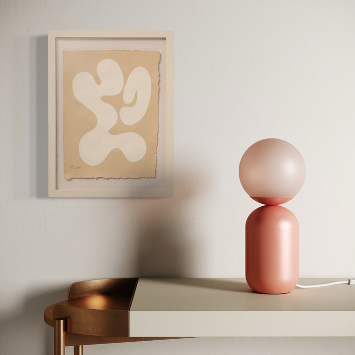 The artwork 'Serenity I' by Shadia Sabagh features an abstract organic form in white against a beige background, displayed in a white frame. Alongside, a modern pink spherical table lamp rests on a light-colored surface, with a copper-toned chair visible in the composition. Acrylic on handmade cotton paper.