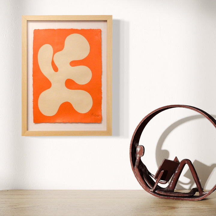 The artwork 'Stillness I' by Shadia Sabagh features an abstract organic shape in cream against a vibrant orange background. The composition presents a flowing, biomorphic form reminiscent of Matisse's cut-outs, painted in acrylic on handmade cotton paper with deckled edges, displayed in a natural wooden frame. A minimalist yet expressive piece that captures motion in stillness.