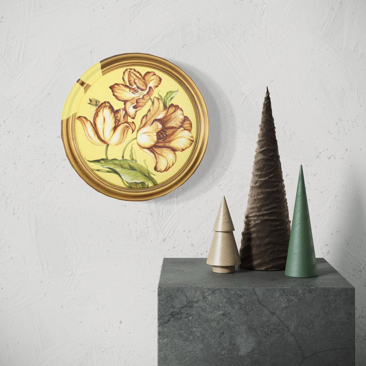 The artwork 'Sunset III' by Carlos Gamez de Francisco features a circular golden frame containing delicate peach-colored tulips with brown accents against a warm yellow background. The floral design showcases elegant brushwork in acrylic on canvas, displayed alongside minimalist cone-shaped decorative objects in muted tones of brown, beige, and sage green on a concrete surface.