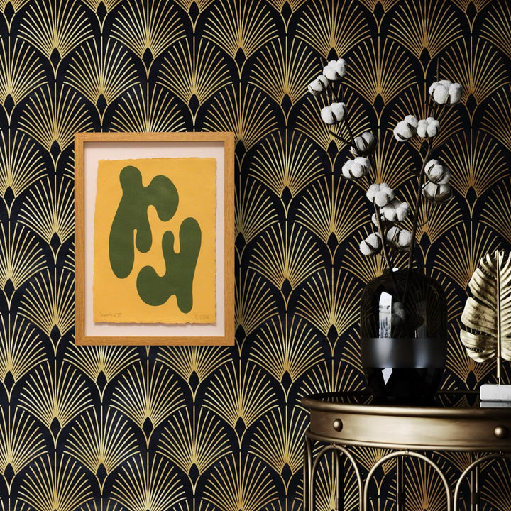 The artwork 'Synergy III' by Shadia Sabagh features abstract olive-green organic shapes on a golden yellow background, framed in white, displayed against art deco-inspired black and gold fan-pattern wallpaper. A black vase with cotton stems and a decorative golden palm leaf complete the modern artistic composition.