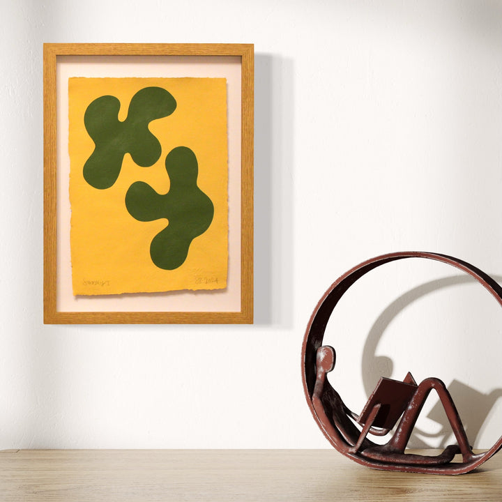 The artwork 'Synergy I' by Shadia Sabagh features two organic, amoeba-like shapes in deep olive green against a vibrant yellow background, framed in natural wood. Created with acrylic paint on handmade cotton paper, the abstract composition suggests fluid movement and natural forms, displaying a minimalist modern aesthetic that emphasizes shape and color contrast. By Shadia Sabagh.