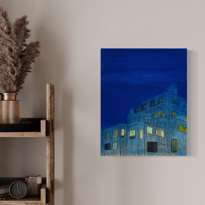 The artwork 'The Blue Hour (enveloping)' features an architectural cityscape painting against a deep cobalt blue twilight sky. An urban building is rendered in muted grays and illuminated yellows, suggesting lit windows at dusk. The oil on linen creates a textural, atmospheric effect capturing the transitional moment between day and night. By Cora Jane Glasser.