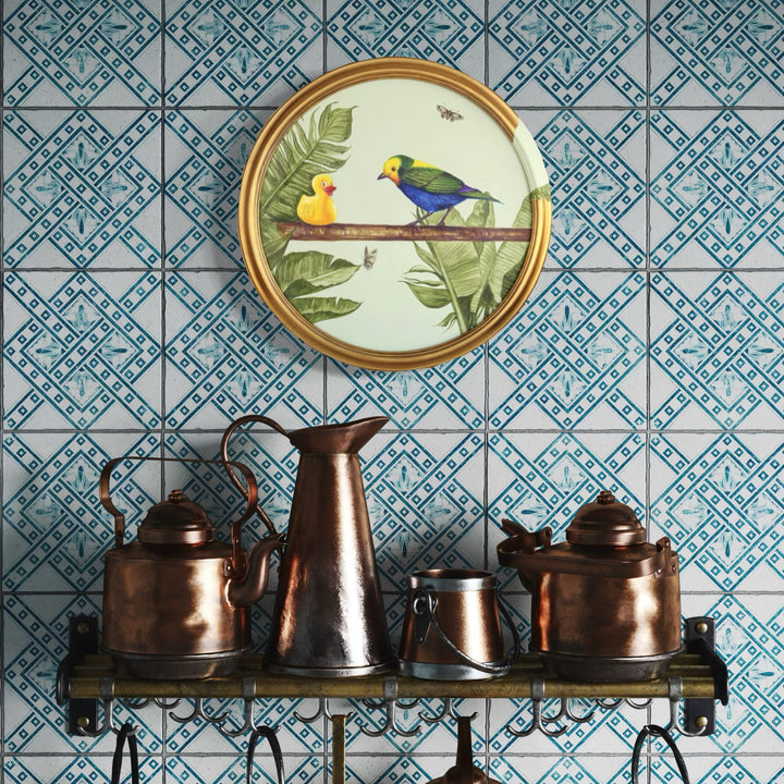 The artwork 'The Conversation' features a circular framed illustration of two birds, one yellow and one blue, perched on a branch against tropical green leaves, mounted on a blue and white geometric patterned wallpaper above vintage copper kettles and pitchers - by Carlos Gamez de Francisco