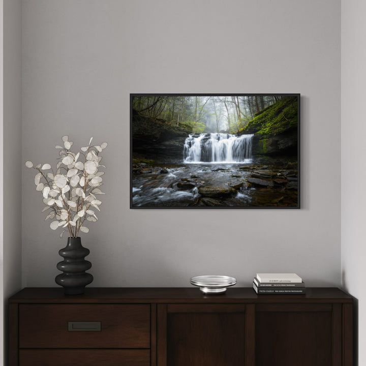 The artwork 'The Music of Water' by Dennis Maida captures a serene waterfall cascading over rocky ledges in a misty forest setting. The photograph features multiple water streams flowing over dark, moss-covered rocks, surrounded by lush green foliage and fog-laden trees. The composition blends dynamic water movement with tranquil forest atmosphere in a fine art nature photograph at 24x36 inches.