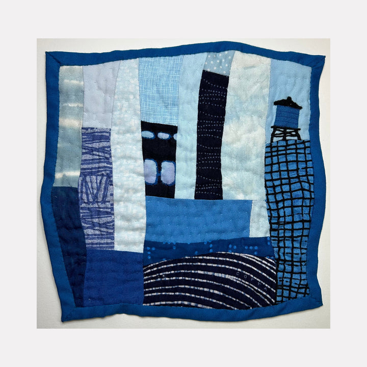 The artwork 'Cityscape (Shades of Blue)' by Ann Cofta features an abstract urban landscape quilt in various blue tones. Geometric shapes suggest buildings and a water tower, created through hand quilting and appliqué techniques. The patchwork design combines textured fabrics with embroidered details, bordered by a deep blue frame, creating a modern interpretation of a city skyline.