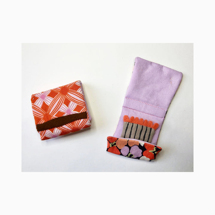 The artwork 'Matchbooks (Lilac & Orange)' by Ann Cofta features two fabric matchbooks - one with a vibrant orange and white geometric pattern, and another in soft lilac with decorative hand-sewn matchsticks and floral embellishments. Each piece showcases detailed applique work, combining fabric and paint in a modern textile interpretation of everyday objects. - Ann Cofta